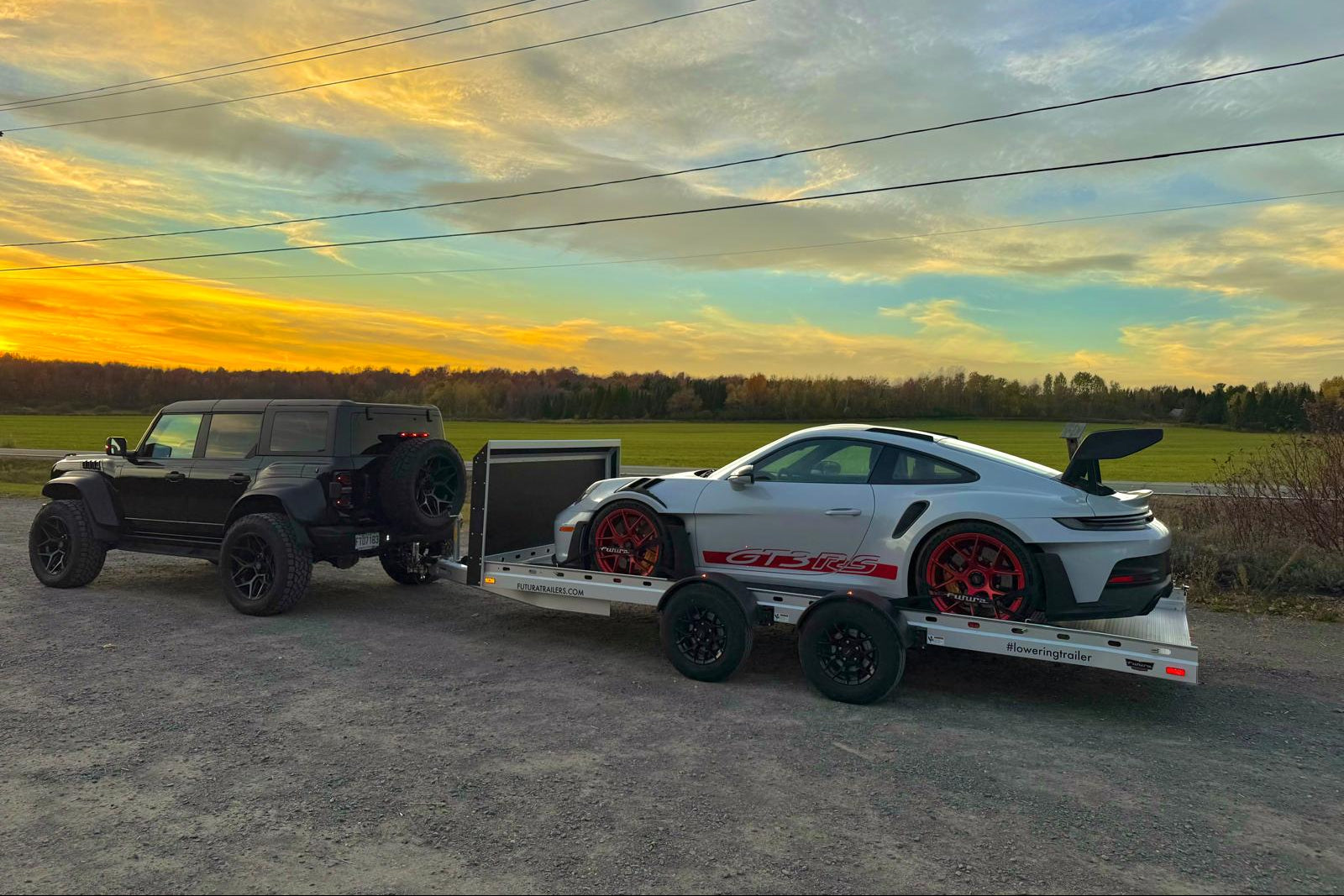 Black SUV towing a trailer with a white sports car against a sunset sky.